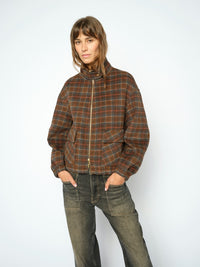 A woman modeling a Mos Mosh brown and orange plaid zip-up jacket with a high collar, paired with dark grey jeans, against a plain white studio background.