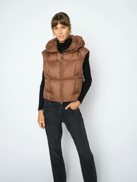 A woman modeling a brown hooded Mos Mosh puffer vest over a black turtleneck and black jeans, standing against a light gray studio background.