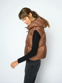 A woman wearing a brown Mos Mosh puffer vest over a black long-sleeve top and dark jeans, photographed against a plain light gray studio background.