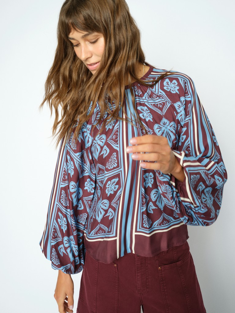 Model wears a long-sleeve blouse with a blue and burgundy floral block print, paired with burgundy pants, against a plain light grey background.