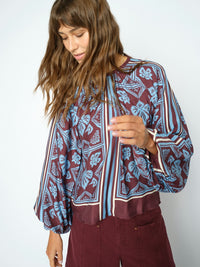 Model wears a long-sleeve blouse with a blue and burgundy floral block print, paired with burgundy pants, against a plain light grey background.