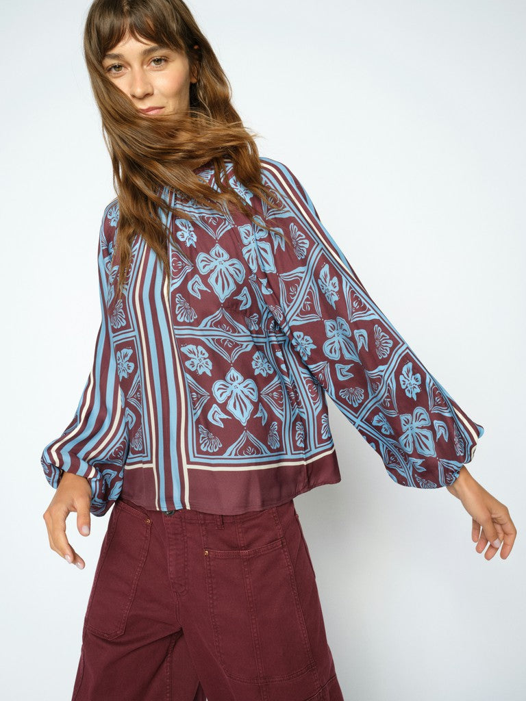 Model wears a long-sleeve blouse with a blue and burgundy floral block print, paired with burgundy pants, against a plain light grey background.