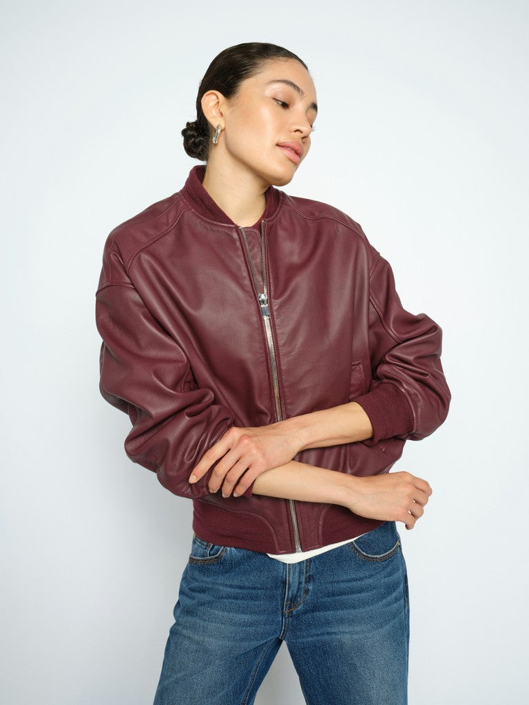 Model wears a Mos Mosh burgundy leather bomber jacket and blue jeans, standing against a light grey studio background.