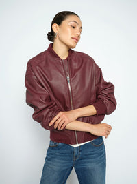 Model wears a Mos Mosh burgundy leather bomber jacket and blue jeans, standing against a light grey studio background.