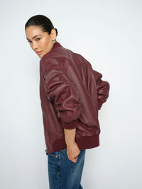Model wears a burgundy Mos Mosh leather bomber jacket and blue jeans, photographed from the back-three-quarter angle against a plain light gray studio background.