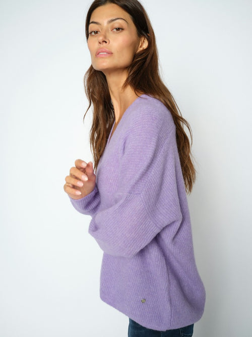 Model wears a Mos Mosh Thora knit sweater with a deep V-neck and long sleeves in lilac, paired with dark blue jeans, standing against a plain white background.