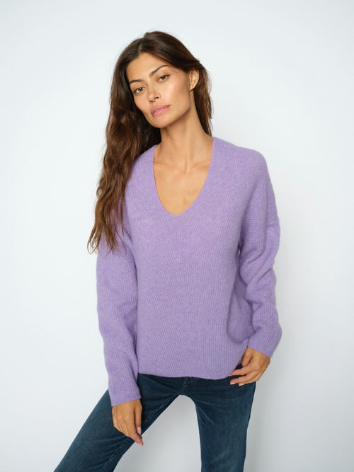 Model wears a Mos Mosh Thora knit sweater with a deep V-neck and long sleeves in lilac, paired with dark blue jeans, standing against a plain white background.