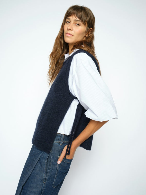 Model wears a dark navy blue Mos Mosh knit sweater vest over a white short-sleeve button-up shirt and dark blue wide-leg trousers