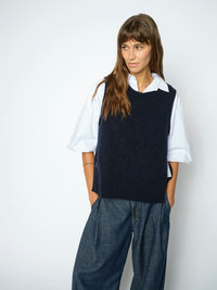 Model wears a dark navy blue Mos Mosh knit sweater vest over a white short-sleeve button-up shirt and dark blue wide-leg trousers