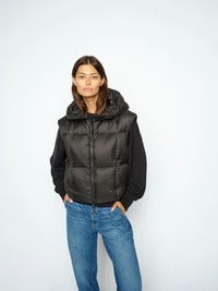 A model modeling a black Mos Mosh hooded puffer vest over a black long-sleeve top and blue jeans, standing against a plain light grey studio background.