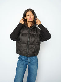 A model wearing a black Mos Mosh puffer vest over a black long-sleeve top and blue jeans, standing against a plain light gray background.