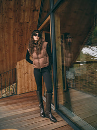 A model stands on a wooden deck in front of a modern wooden building with large glass windows. She is wearing a Mos Mosh tan quilted puffer vest over a black long-sleeve top, black pants, and dark brown knee-high boots.