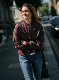 Model standing on a city street wearing a dark burgundy Mos Mosh leather bomber jacket and blue jeans, with her arms crossed in front of her.