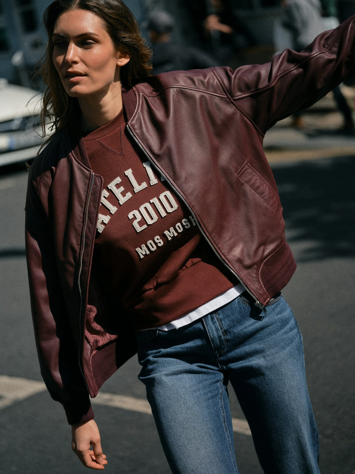 Model standing on a city street wearing a dark burgundy Mos Mosh leather bomber jacket and blue jeans