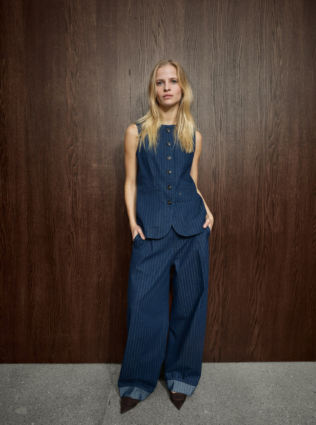 A model stands against a dark wood-paneled wall wearing a matching sleeveless button-front vest and wide-leg trousers set in dark blue pinstriped fabric, paired with dark pointed shoes.