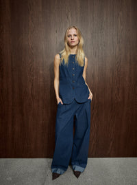 A model stands against a dark wood-paneled wall wearing a matching sleeveless button-front vest and wide-leg trousers set in dark blue pinstriped fabric, paired with dark pointed shoes.