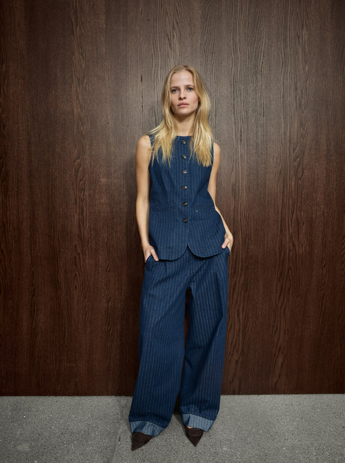 A model stands against a dark wood-paneled wall wearing a matching sleeveless button-front vest and wide-leg trousers set in dark blue pinstriped fabric, paired with dark pointed shoes.