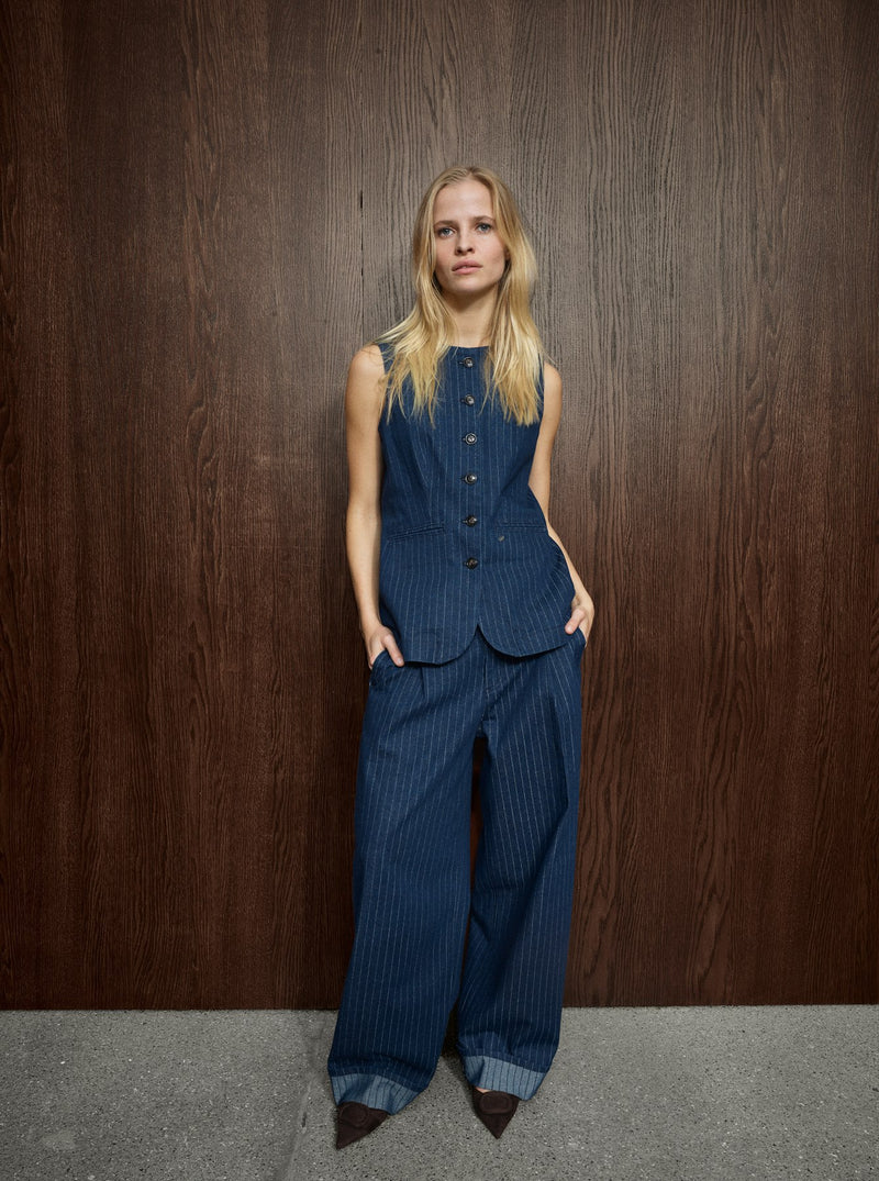 A model stands against a dark wood-paneled wall wearing a matching sleeveless button-front vest and wide-leg trousers set in dark blue pinstriped fabric, paired with dark pointed shoes.