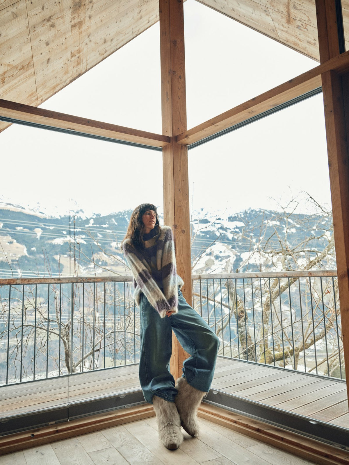 Model wears a striped Mos Mosh sweater and jeans sitting indoors with a view of a cityscape through large windows.