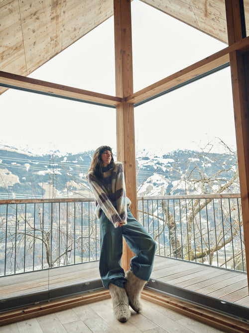Model wears a striped Mos Mosh sweater and jeans sitting indoors with a view of a cityscape through large windows.
