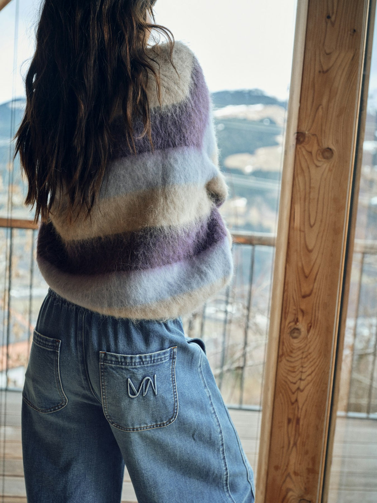 Model wears a striped Mos Mosh sweater and jeans sitting indoors with a view of a cityscape through large windows.
