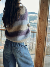 Model wears a striped Mos Mosh sweater and jeans sitting indoors with a view of a cityscape through large windows.
