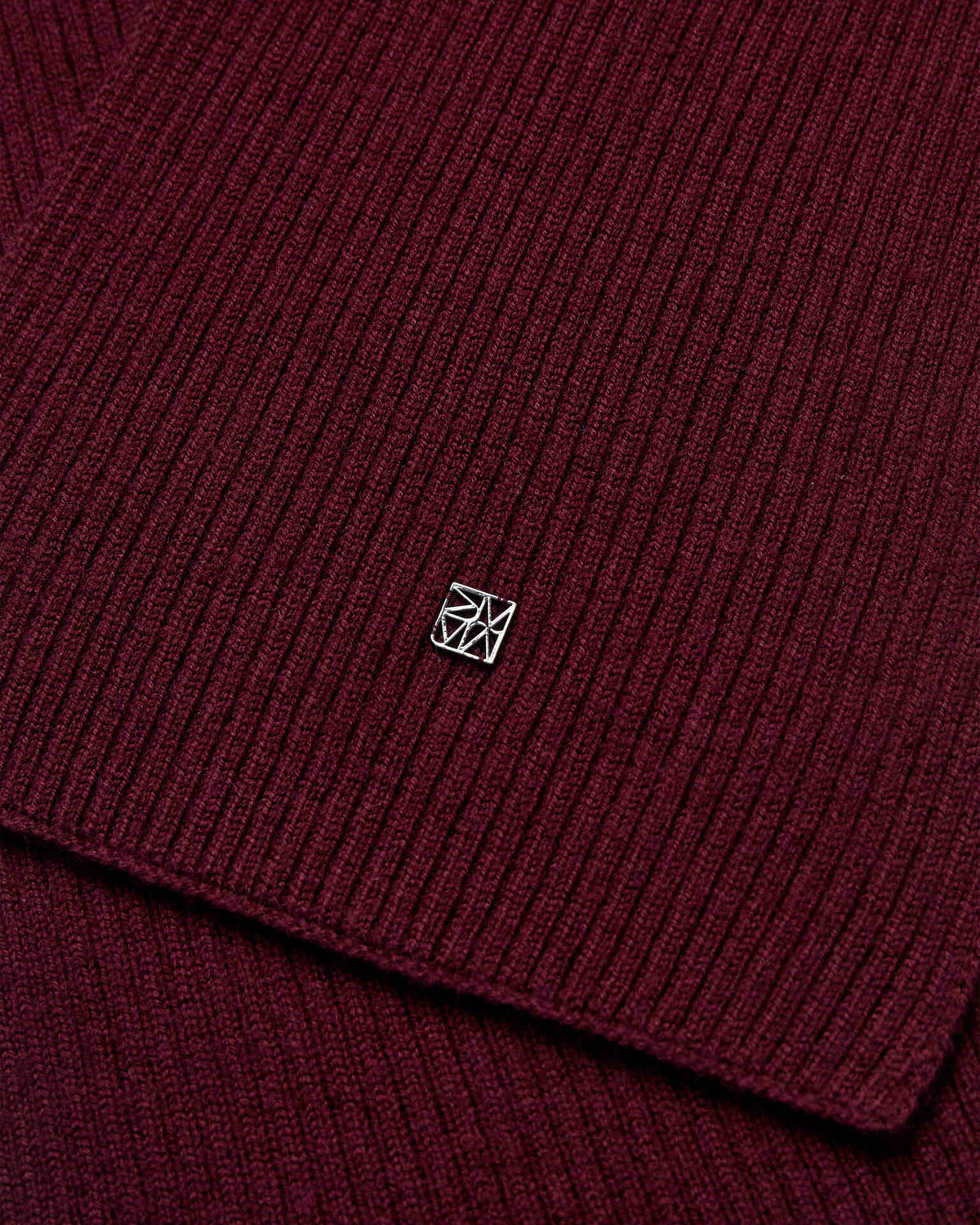 Close-up of the dark burgundy ribbed wool knit Mos Mosh Scarf with a small logo charm in the centre