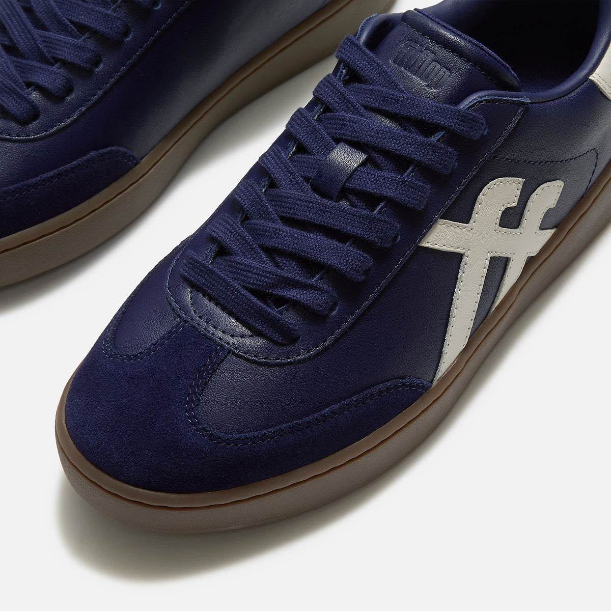 A close-up of FitFlop Dash navy blue suede and leather sneaker with matching navy laces, a white side stripe, and a brown gum sole, available at The hoe Gallery