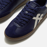 A close-up of FitFlop Dash navy blue suede and leather sneaker with matching navy laces, a white side stripe, and a brown gum sole, available at The hoe Gallery