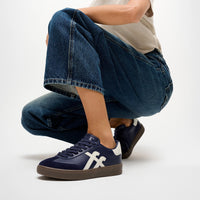 Model wears FitFlop Dash navy blue suede and leather sneaker with matching navy laces, a white side stripe, and a brown gum sole, available at The hoe Gallery