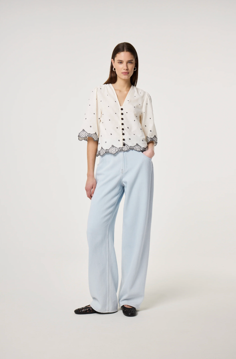 Model wears the Fabienne Chapot Tylera Blouse in ecru with small black polka dots and edged with embroidered fish at both the hemline and the three quarter sleeve edge. It has tiny black button central closure