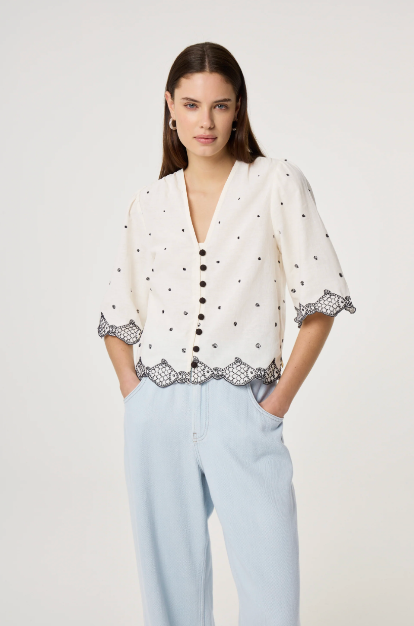 Model wears the Fabienne Chapot Tylera Blouse in ecru with small black polka dots and edged with embroidered fish at both the hemline and the three quarter sleeve edge. It has tiny black button central closure