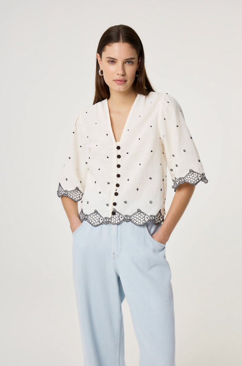 Model wears the Fabienne Chapot Tylera Blouse in ecru with small black polka dots and edged with embroidered fish at both the hemline and the three quarter sleeve edge. It has tiny black button central closure